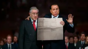 Due uomini in giacca e cravatta mostrano un foglio davanti a un pubblico.