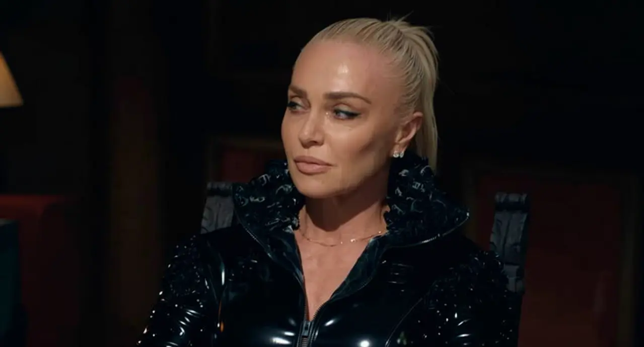 Una donna con capelli biondi legati e giacca nera lucida seduta in un ambiente poco illuminato