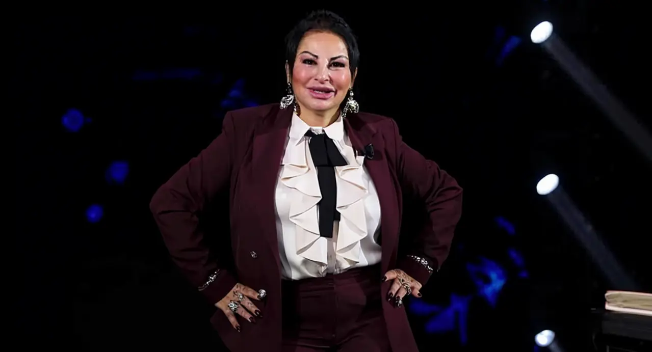 Una donna in abito elegante posa su un set televisivo con sfondo scuro e luci