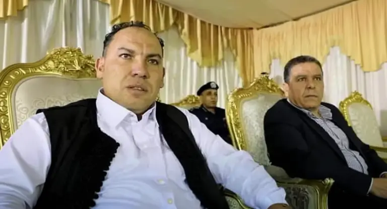 Due uomini seduti su poltrone dorate in un ambiente formale con una guardia sullo sfondo