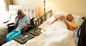 Due anziani in una stanza, uno a letto e l'altra in sedia a rotelle, circondati da targhe commemorative