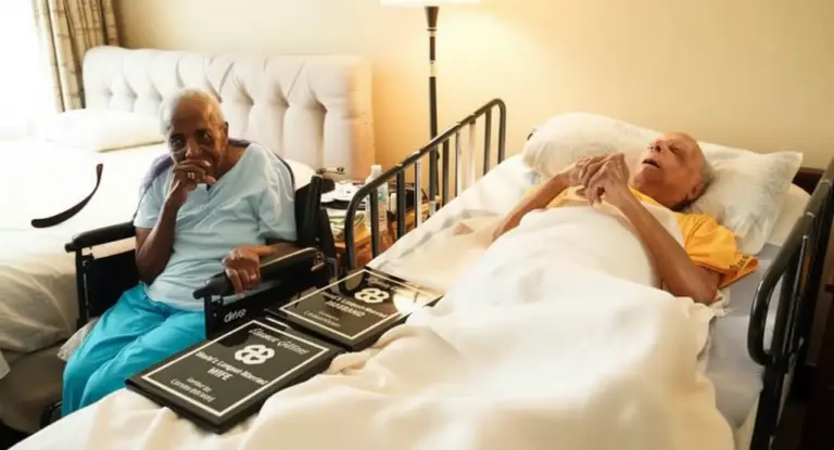 Due anziani in una stanza, uno a letto e l'altra in sedia a rotelle, circondati da targhe commemorative