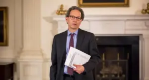 Un uomo in abito formale tiene dei documenti in una sala elegante con camino.