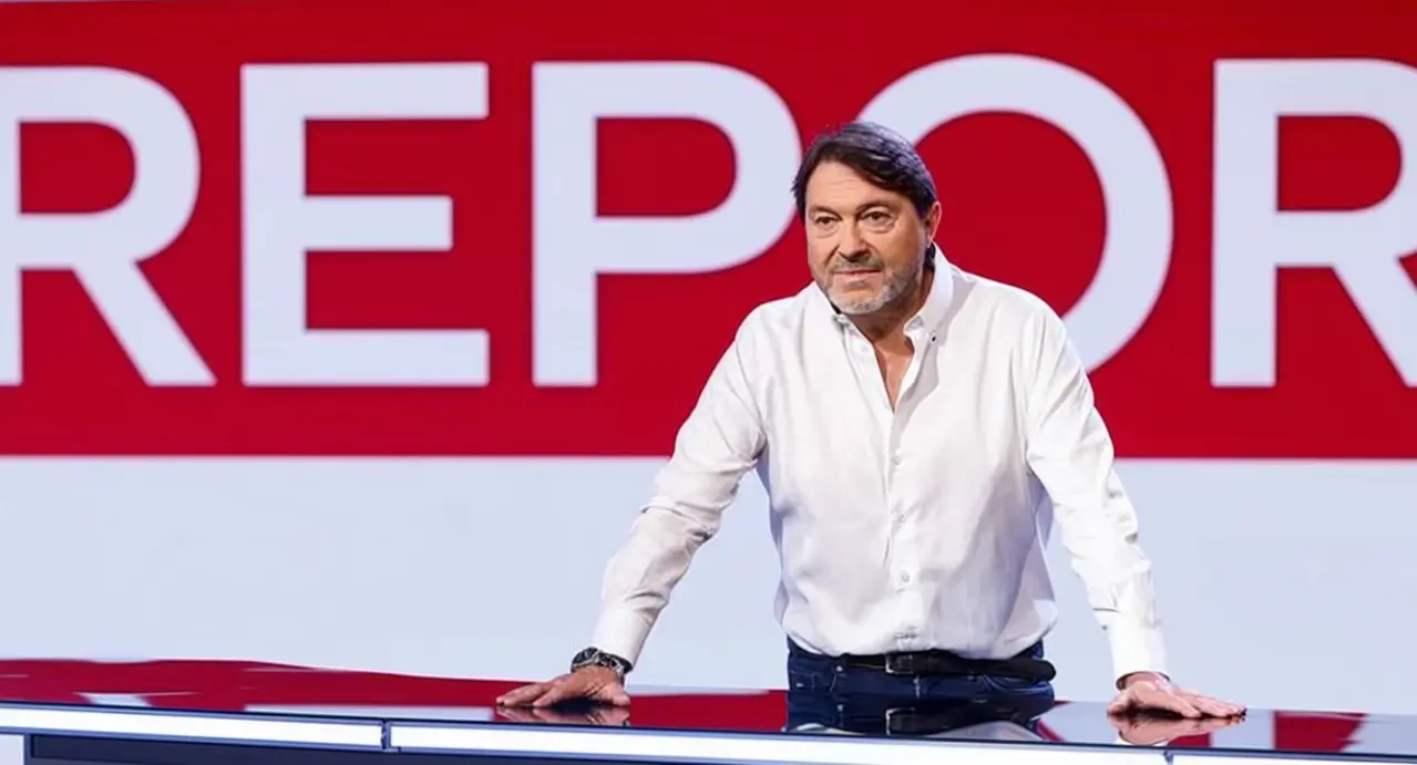 Un uomo in camicia bianca appoggiato a un tavolo, davanti a uno sfondo rosso con scritta parziale.